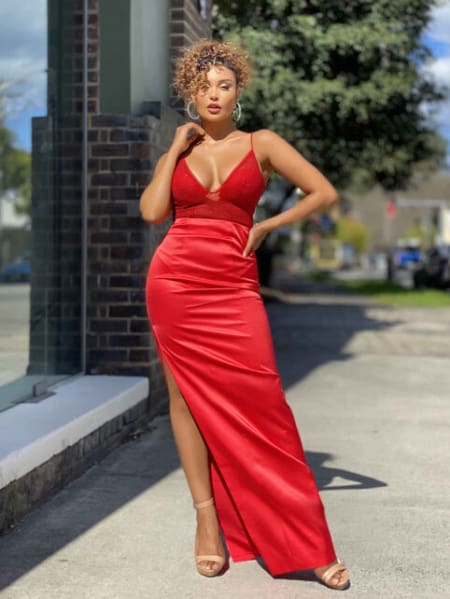 A woman stands confidently outdoors, embodying a true DIVA in her vibrant, red sleeveless gown with a high slit. She has curly hair, large earrings, and open-toed high heels. The background features a brick wall, tree, and a suburban street with blurred houses and cars, adding charm to her DIVA JX4061 look. Angels Formal Wear