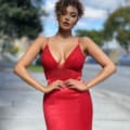 A DIVA JX4061 with curly hair stands confidently outdoors, wearing a form-fitting sleeveless red dress with a deep neckline. Their hands rest on their hips against a blurred background of trees and a cloudy sky. Angels Formal Wear