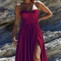 A woman stands against a rocky background, wearing a strapless red gown with a sweetheart neckline and a thigh-high slit. She has shoulder-length wavy hair and is accessorized with small earrings. The elegance of her attire and the serene confidence in her stance evoke the allure of DAPHNE PO927. Angels Formal Wear