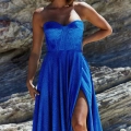 A woman stands in front of a rocky background wearing a vibrant blue strapless gown. The DAPHNE PO927 dress has a sweetheart neckline, fitted bodice, and a high side slit, revealing one leg. She poses with one hand on her hip and wears elegant earrings. Angels Formal Wear