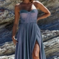 A woman in a strapless, shimmering gown with a high slit poses confidently in front of a rocky background that could be straight out of DAPHNE's world. She has short, wavy hair and is wearing drop earrings. The dress, DAPHNE PO927, has a structured bodice and flows elegantly to the ground. Angels Formal Wear