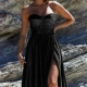 A woman stands outdoors against a rocky background, wearing a strapless black evening gown with a structured bodice and a high slit on one side. She has shoulder-length hair and is accessorized with DAPHNE PO927 earrings, posing in an elegant and confident manner reminiscent of Daphne's grace. Angels Formal Wear