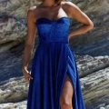 A woman stands against a rocky background, wearing a strapless, blue DAPHNE PO927 dress with a high slit. She has shoulder-length, wavy hair and poses with one hand on her hip. She accessorizes with drop earrings and exudes confidence in a stylish, elegant look by DAPHNE. Angels Formal Wear