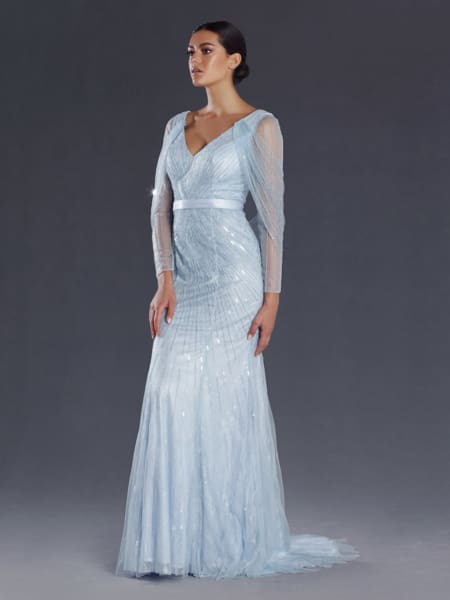 A woman stands against a gray background wearing the ABRIEL JX101, an elegant light blue gown with sheer long sleeves and a V-neckline. The dress has a fitted bodice and flares slightly at the bottom. Her hair is styled in an updo, and she gazes to the side. Angels Formal Wear