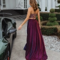 A woman in a vibrant, flowing gown with a detailed bodice is walking away from the camera towards a large, elegant building in Manchester. The gown, adorned in shades of purple, red, and blue, complements the luxurious green car and neatly trimmed topiary nearby. MANCHESTER PO893 Angels Formal Wear