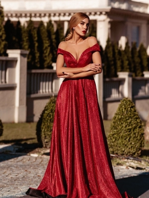 A woman stands outdoors near a building in an elegant, off-the-shoulder, red gown. The dress is floor-length and made of a shimmering fabric. She has long, wavy hair and is posing with her arms relaxed at her sides, looking off into the distance, exuding grace reminiscent of an EMPRESS TW028. Angels Formal Wear