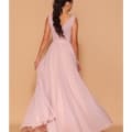 A woman with long, dark hair in a ponytail is shown from behind, wearing a flowing, sleeveless pale pink ELIZA LD1105 gown from Formal Wear Shop Sunshine Coast. Standing against a light brown background and slightly lifting her dress to the side, she creates a sense of movement. Angels Formal Wear