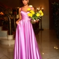 A woman in an elegant, floor-length, pink satin EMMY BB210 stands in a luxurious setting reminiscent of a Formal Wear Shop on the Sunshine Coast. She holds a bouquet of yellow flowers in one hand and has the other hand in the pocket of her dress. The background features a grand staircase and warm lighting. Angels Formal Wear