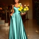 A woman in an elegant teal satin gown stands in a sophisticated indoor setting, reminiscent of EMMY BB210 on Sunshine Coast. She holds a bouquet of yellow flowers in one hand and has the other hand in her dress pocket. The background features a grand staircase and ornate decor. Angels Formal Wear