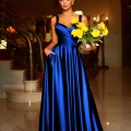 A woman in a long, elegant blue EMMY BB210 satin gown stands indoors holding a bouquet of yellow flowers. She has her hands in the dress’s pockets and is positioned in front of a staircase with ornate railings. The background features warm lighting from chandeliers. Angels Formal Wear