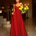 A woman in a flowing red evening dress stands in an elegant room, holding a glass bowl of yellow flowers. The room features a grand staircase with ornate railings and warm lighting from chandeliers above, creating a setting worthy of an EMMY BB210. Angels Formal Wear