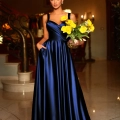 A woman in an elegant, floor-length blue satin gown stands on a polished marble floor in a luxurious interior, holding a vase of yellow flowers. The background includes ornate railings, warm lighting, and a staircase, with the sophisticated ambiance only rivaled by her stunning EMMY BB210 heels. Angels Formal Wear