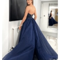 A woman in a strapless, navy blue evening gown, the EVE JX6006, with a flowing skirt poses in a modern, white room. She looks over her shoulder toward the camera, revealing the dress's detailed bodice and open back. A large mirror in the background reflects her full image. Angels Formal Wear