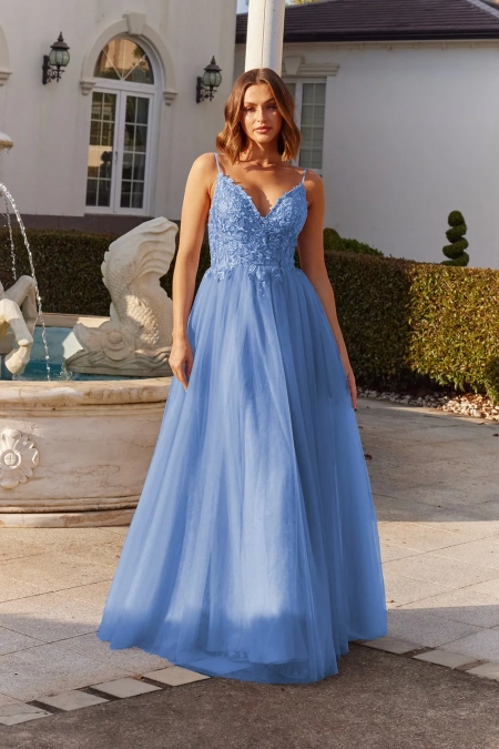 A woman is standing outdoors near a decorative fountain, wearing a long, flowing, light blue gown with a lace bodice and thin straps. The background includes trimmed hedges and parts of what seems to be the classical-style ELSIE PO984. Angels Formal Wear