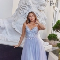 A woman in a lavender, embellished gown stands in front of a large white lion statue, labeled ELSIE PO984. She has one hand resting on the statue's pedestal and is smiling slightly. The background features an ornate building with arched windows and greenery. Angels Formal Wear