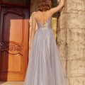 A woman in a flowing, floor-length silver gown with a low back and delicate straps, labeled ELSIE PO984, stands with her back facing the camera. She has one hand resting on a stone column. The backdrop features a wooden door with ornate carvings. Sunlight illuminates her and the setting. Angels Formal Wear