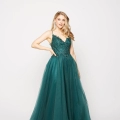 A woman stands against a plain white background wearing a sleeveless, emerald green ball gown. The ELSIE PO984 dress features a fitted, embellished bodice with delicate floral designs and a voluminous tulle skirt. She has long, wavy blonde hair and one hand resting on her hip. Angels Formal Wear