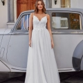 A woman in a white sleeveless wedding dress stands in front of a vintage car. The dress, labeled ELSIE PO984, has a lace bodice and a flowing skirt. She is posing in front of an ornate double door with pillars and lanterns, suggesting a formal or elegant setting. Her radiant smile reveals her joy on this special day. Angels Formal Wear