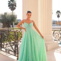 A woman stands on a sunlit balcony, wearing a vibrant turquoise ball gown with delicate lace detailing from the ELICIA TY300. The balcony features ornate railings and columns, while palm trees sway in the background. She has her hair styled up and smiles softly at the camera. Angels Formal Wear