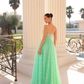 A woman in a pastel green, sleeveless ELICIA TY300 stands in front of tall white columns, looking over her shoulder. The dress features a fitted bodice and a full, textured skirt. She is on an elegant balcony with ornate railings. Palm trees and a clear sky are visible in the background. Angels Formal Wear