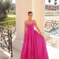 A woman stands on a sunlit balcony, in front of a large white pillar. She is wearing a bright pink, floral-textured gown with a fitted bodice and flowing skirt by ELICIA TY300. The background features ornate railings, a swimming pool, palm trees, and distant scenery. Angels Formal Wear