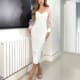 A woman stands in a sleek, white room wearing a fitted ADORE JX6008 with sheer sleeves and strappy white high heels. She has long hair and poses confidently beside a radiator and a black and white abstract painting on the wall. Angels Formal Wear