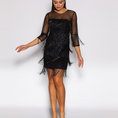 A woman in a black dress with fringe details, reminiscent of the Gatsby era, stands against a plain white background. The GATSBY JX2061 features three-quarter sleeves and a sheer neckline. She is wearing transparent high-heeled shoes, her hair pulled back, and her arms slightly outstretched. Angels Formal Wear