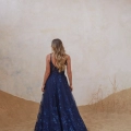 A person with long, wavy hair stands with their back to the camera, wearing a navy blue, floor-length, sleeveless LIR PO2471 with floral patterns. The setting appears to be a sandy, earth-toned environment on the Sunshine Coast, hinting at an elegant dress shop nearby. Angels Formal Wear