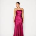 A woman stands against a plain background wearing a strapless, floor-length deep magenta satin MOSELLE PO2457 with a fitted bodice and a subtle mermaid-style skirt, perfect for any Dress Shop on the Sunshine Coast. She has her hair styled in a slicked-back updo and is accessorizing with dangling earrings. Angels Formal Wear