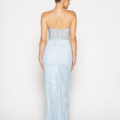 A woman is shown from the back, wearing the ALMA PO2441 dress. This light blue, strapless dress features a fitted bodice with sheer panel detailing and flows into a long, straight skirt. Her hair is styled in a neat, low bun against a plain white background. Angels Formal Wear