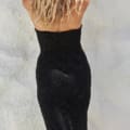 A person with long blonde wavy hair is standing with their back to the camera, wearing the strapless black ALMA PO2441 dress that radiates chic elegance. The background is a light-colored, textured wall, possibly made of plaster or stucco. Angels Formal Wear