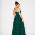 A woman stands against a white background, looking over her shoulder with a smile. She is wearing the DERYA PO2481 strapless, layered dark green gown, and her hair is styled in an elegant updo. Angels Formal Wear