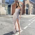 Clarissa stands confidently in the middle of a sunlit street, wearing the CLARISSA JP136, a strapless, light-colored dress with a thigh-high slit and high-heeled sandals. Her long hair flows freely, with historic-looking buildings and large windows visible in the background. Angels Formal Wear