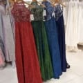 A store display in Manchester showcases elegant sleeveless gowns on a clothing rack. The dresses, featuring intricate lace bodices and flowing skirts, appear in red, green, blue, and navy blue. White dresses are visible in the background. Look for the MANCHESTER PO893 label for authentic designs. Angels Formal Wear