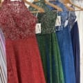 Four elegant evening gowns are displayed on wooden hangers in our Manchester boutique. The MANCHESTER PO893 collection features dresses in red, green, blue, and dark blue, all adorned with intricate silver sequin patterns on the bodice. Each has a flowing, sparkly skirt and tags hanging from the neckline. Angels Formal Wear