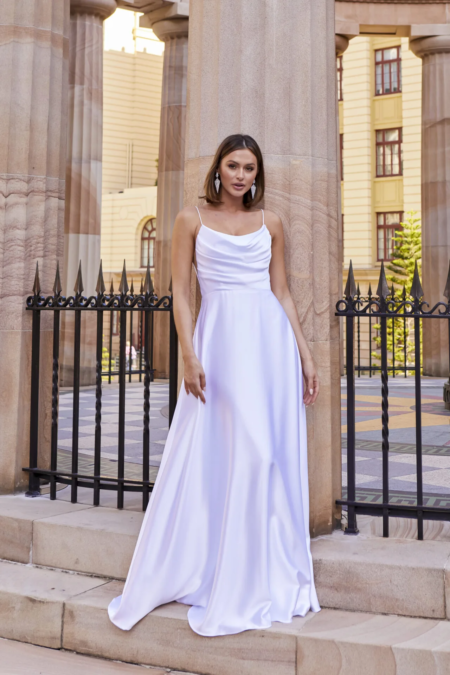 A woman in a long, flowing white dress stands in front of tall stone columns, reminiscent of an ESTHER PO944-like elegance. She has short, brown hair and is wearing dangling earrings. The background features a wrought iron fence and a multi-story building with large windows and yellow walls. Angels Formal Wear