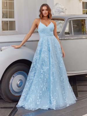 A woman wearing a light blue, floor-length, lace gown stands beside a vintage silver car. The BRIONY PO972 dress features a fitted bodice with spaghetti straps and a voluminous skirt. She is leaning gracefully against the car, which is parked in front of a white building. Angels Formal Wear