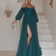 A woman stands on a staircase at a Sunshine Coast dress shop, wearing an elegant LILY PO2305 with sheer long sleeves and a high slit revealing her leg. Her hair is styled elegantly, complemented by minimal accessories. The background is simple, featuring candles on the steps. Angels Formal Wear