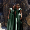 Two women stand closely together on a sandy beach in front of large rocks, each wearing shiny, dark green, floor-length dresses with short, ruffled sleeves. With relaxed expressions and natural hairstyles, they exude elegance reminiscent of PETAL TO882 as one woman slightly tilts her head. Angels Formal Wear