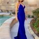 A woman in a vibrant blue, floor-length gown with a deep neckline stands beside a small pool. She is on a stone pathway bordered by HAZEL PO815 flower beds, and the background features a building with a stone facade. She has a poised and confident expression. Angels Formal Wear