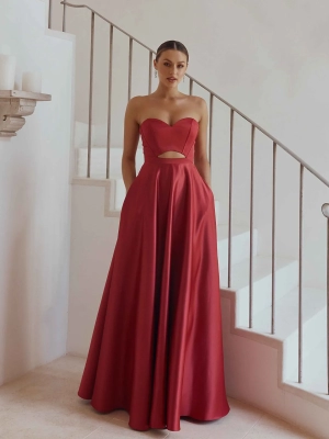 A woman stands on an elegant staircase, wearing a strapless, floor-length red gown with a cutout midriff. She is posing with her hands in the gown's pockets, her hair pulled back and minimal makeup complete the look. White walls and a small wall fixture with candles are in the background. The sophisticated attire epitomizes HOLLIE PO2313. Angels Formal Wear