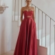 A woman stands on an elegant staircase, wearing a strapless, floor-length red gown with a cutout midriff. She is posing with her hands in the gown's pockets, her hair pulled back and minimal makeup complete the look. White walls and a small wall fixture with candles are in the background. The sophisticated attire epitomizes HOLLIE PO2313. Angels Formal Wear