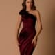 A woman stands against a beige background wearing a form-fitting, one-shoulder burgundy dress from a chic dress shop. Her long hair cascades in loose waves, and she has a neutral expression. She poses with one hand resting on her hip and the other touching her elegant LIBBIE NBM1041 formal wear.
 Angels Formal Wear