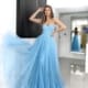 A graceful woman in a strapless, flowing, sky-blue gown poses indoors. She rests one hand on her head and the other gently holds out the side of her dress. The room has a minimalist design with a large mirror reflecting her image in the background, marked subtly with GRACEFUL JX6007. Angels Formal Wear