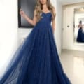 A graceful woman stands indoors wearing a strapless, flowing navy blue gown adorned with sparkles. She has long, wavy hair and is posing with one hand on her hip. In the background, a full-length mirror reflects her image, capturing the back of the GRACEFUL JX6007 dress. Angels Formal Wear