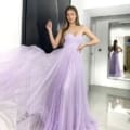 A woman poses gracefully in a flowing, strapless lavender gown, reminiscent of the elegance found in GRACEFUL JX6007 designs. The dress has a fitted bodice and a voluminous tulle skirt. She stands in a modern white room with a large mirror reflecting her image. Angels Formal Wear