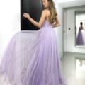 A graceful woman with long hair stands indoors in a flowing, strapless lavender gown. Facing away from the camera, her hand gently touches a black and white wall-mounted artwork. A mirror in the background captures her elegant reflection. The scene exudes an ethereal charm akin to GRACEFUL JX6007. Angels Formal Wear