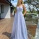 A woman stands on a wooden deck wearing the stunning GLITZ JX5008, a floor-length, strapless, light lavender gown with a sweetheart neckline. She has long, wavy blonde hair and is looking directly at the camera, embodying pure glitz. The background includes a house and some trees. Angels Formal Wear