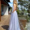 A woman stands on a wooden deck outside a house, posing in a sleeveless, floor-length lavender gown from the GLITZ JX5008 collection. She has long, wavy blonde hair and stands with her hands in the gown's pockets. The background features trees and a partially visible glass railing that adds a touch of glitz. Angels Formal Wear