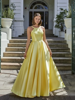 A woman stands outdoors in front of marble steps and an entrance with large white columns. She is wearing a flowing yellow satin gown with a halter neckline and a fitted bodice from the GLISS PO941 collection. Tall potted plants flank her on both sides at the top of the steps. Angels Formal Wear